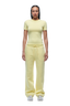 A person with braids and a neutral expression faces forward, wearing the 6PM WMNS T-SHIRT BUTTERCREAM and loose yellow sweatpants with a drawstring, paired with white sneakers.