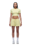 A person with braided hair and a neutral expression faces forward, wearing the 6PM WMNS Cropped T-Shirt Buttercream, matching yellow drawstring shorts, white socks, and white sneakers.