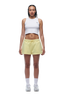 A woman with braided hair and a neutral expression faces forward, wearing a white cropped tank top, 6PM WMNS SHORTS BUTTERCREAM with white side stripes, white socks, and white sneakers.