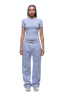 A person stands facing forward, wearing 6PM WMNS SHORTS SOFT VIOLET and a fitted light purple t-shirt with white sneakers. Their hair is braided and they have a neutral expression.
