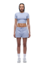 A woman with braided hair and a neutral expression faces forward, wearing the 6PM WMNS T-SHIRT SOFT VIOLET, matching drawstring shorts, white socks, and white sneakers.
