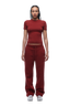 A person with braided hair and a neutral expression faces forward, wearing the 6PM WMNS T-SHIRT WINE RED, matching maroon sweatpants with a drawstring, and white sneakers against a plain white background.