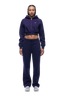 A woman with braided hair and a neutral expression faces forward, wearing the 6PM WMNS Cropped Zip Hoodie in Orbit Blue with matching drawstring sweatpants and white sneakers.
