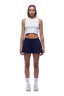 A woman with braided hair and a neutral expression stands facing forward in a white cropped tank, 6PM WMNS SHORTS ORBIT BLUE, white socks, and sneakers against a plain white background.