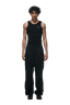 A young man with curly hair faces forward, wearing a black sleeveless tank top, 6PM 6PM+ NYLON PANTS OPEN LEG BLACK with zipped pockets and side piping, and black shoes, against a plain green background.